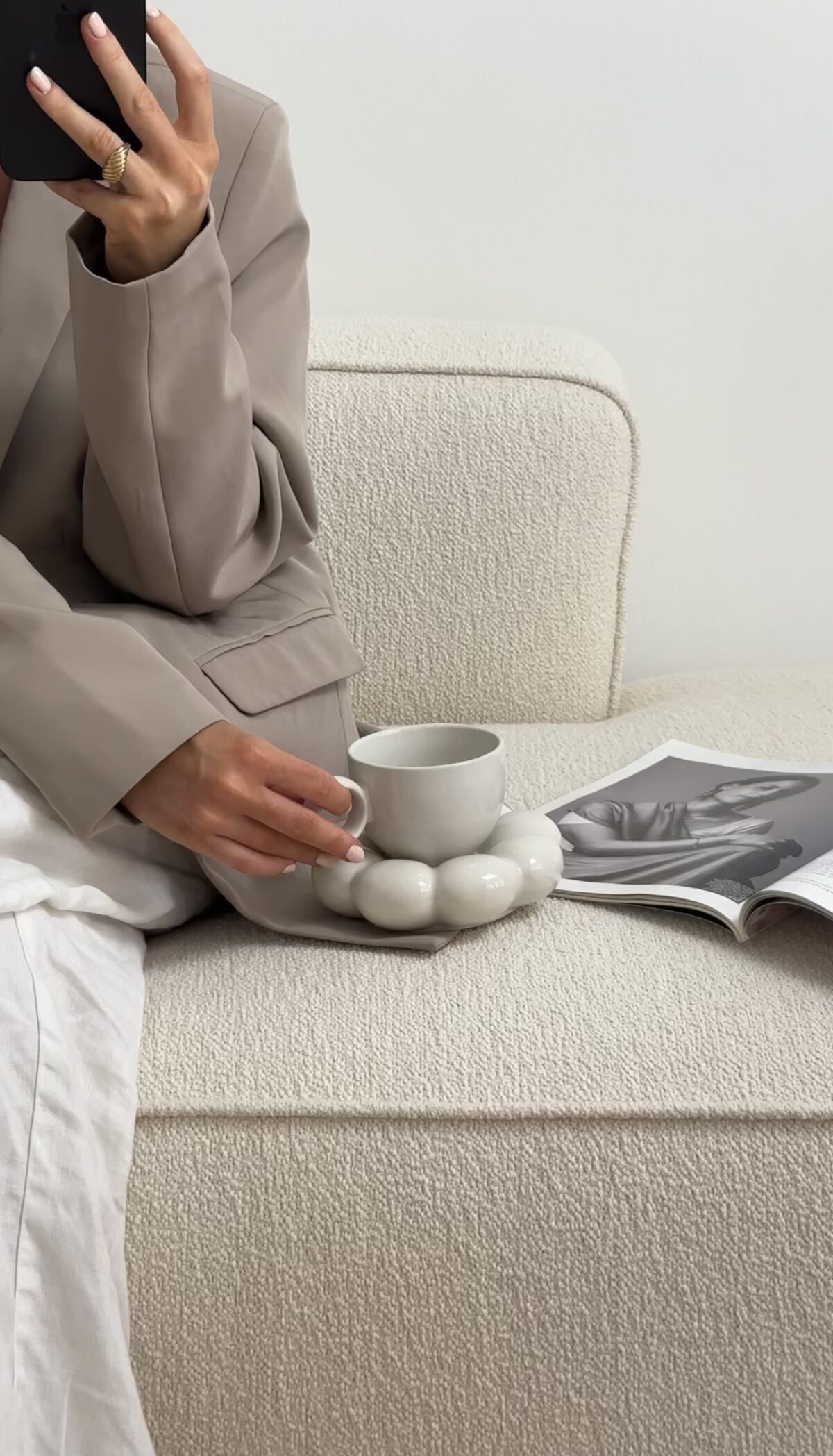 Person in a beige blazer holding a phone, sitting on a sofa with a mug and an open magazine on the seat—perfect for illustrating How to Create Faceless Content.