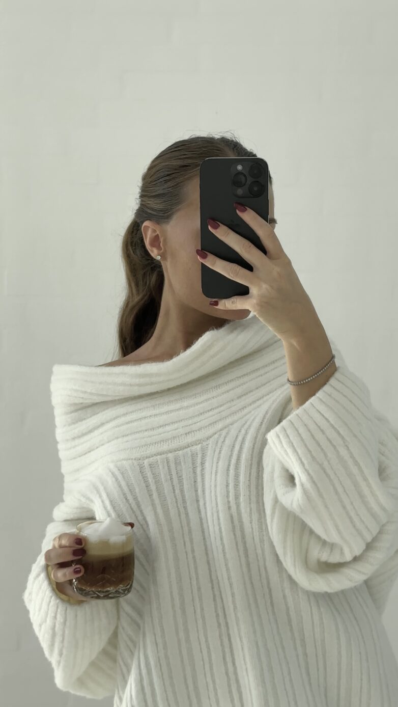 Woman in white sweater takes a mirror selfie holding coffee, nails painted red, hair tied back—an inspiring example of how to create faceless content with style and personality.