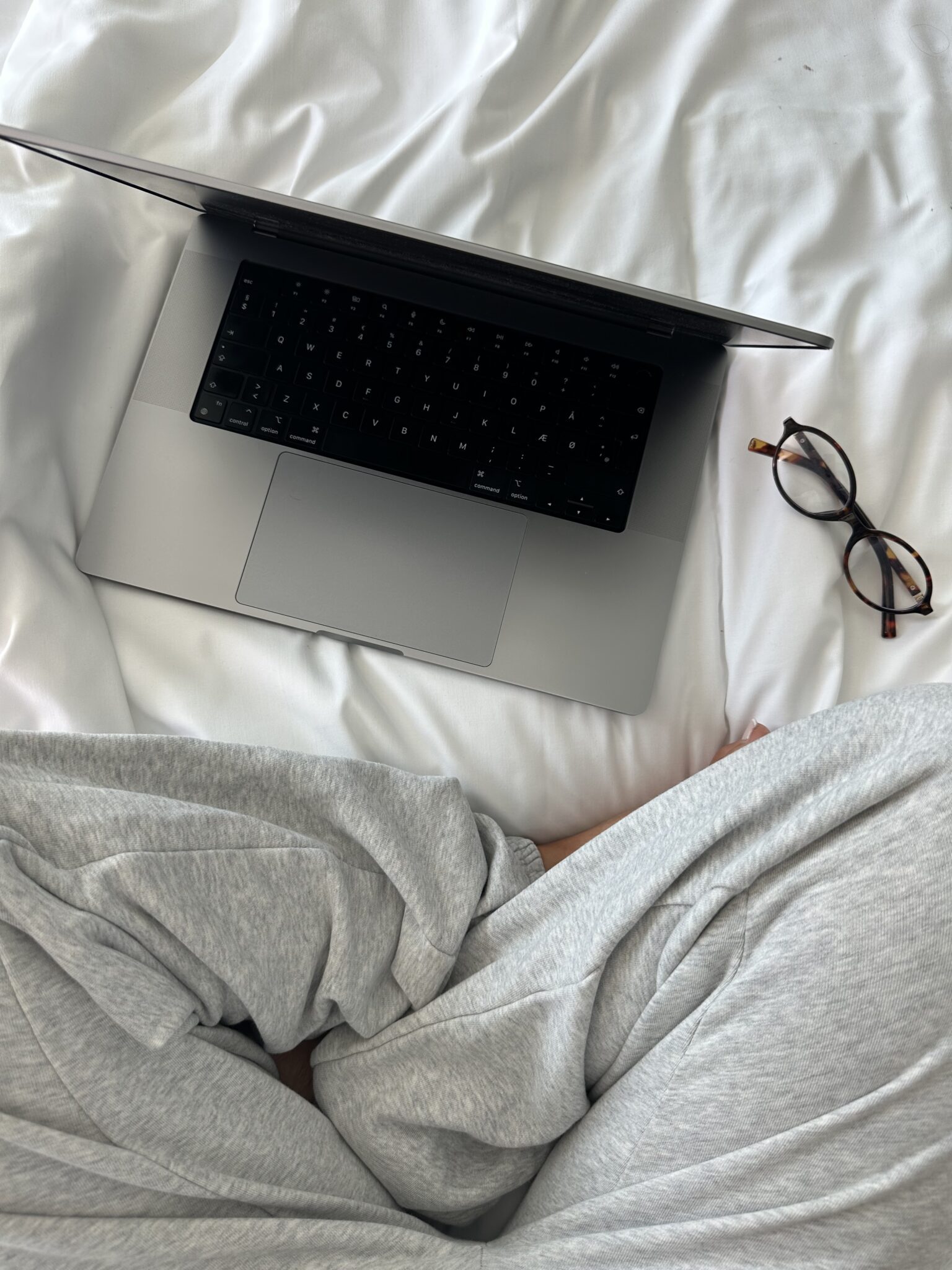 A laptop, eyeglasses, and a person in grey sweatpants sitting cross-legged on a white bed—an ideal scene for building passive income or exploring faceless marketing from the comfort of home.