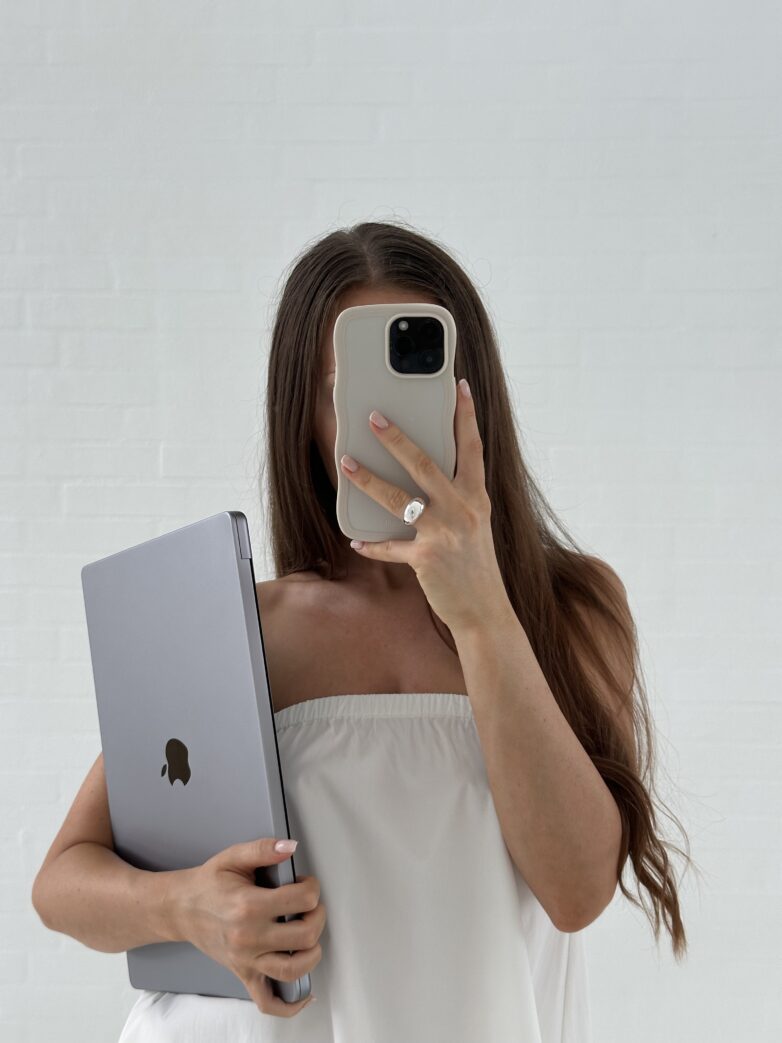 Woman in a white top holding a closed laptop and taking a mirror selfie with her phone—capturing the essence of Faceless Marketing and the freedom that comes with passive income.