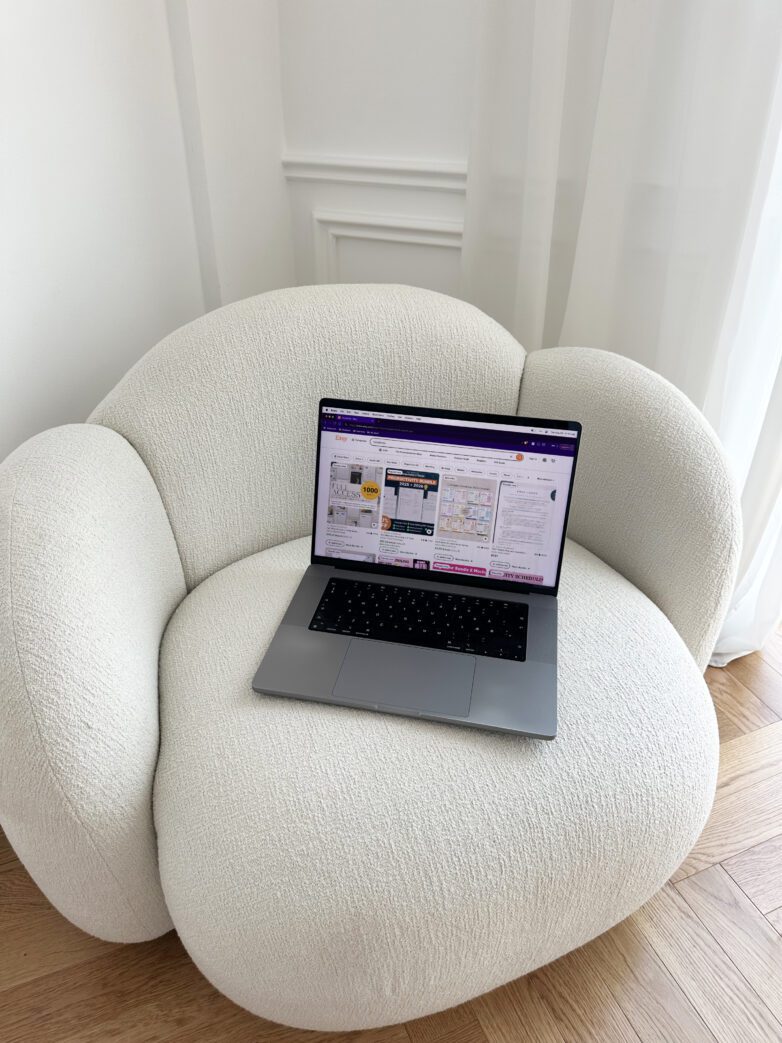 A laptop on a plush, white armchair in a bright, minimalistic room with wooden floors—an inviting workspace for creating faceless digital products.