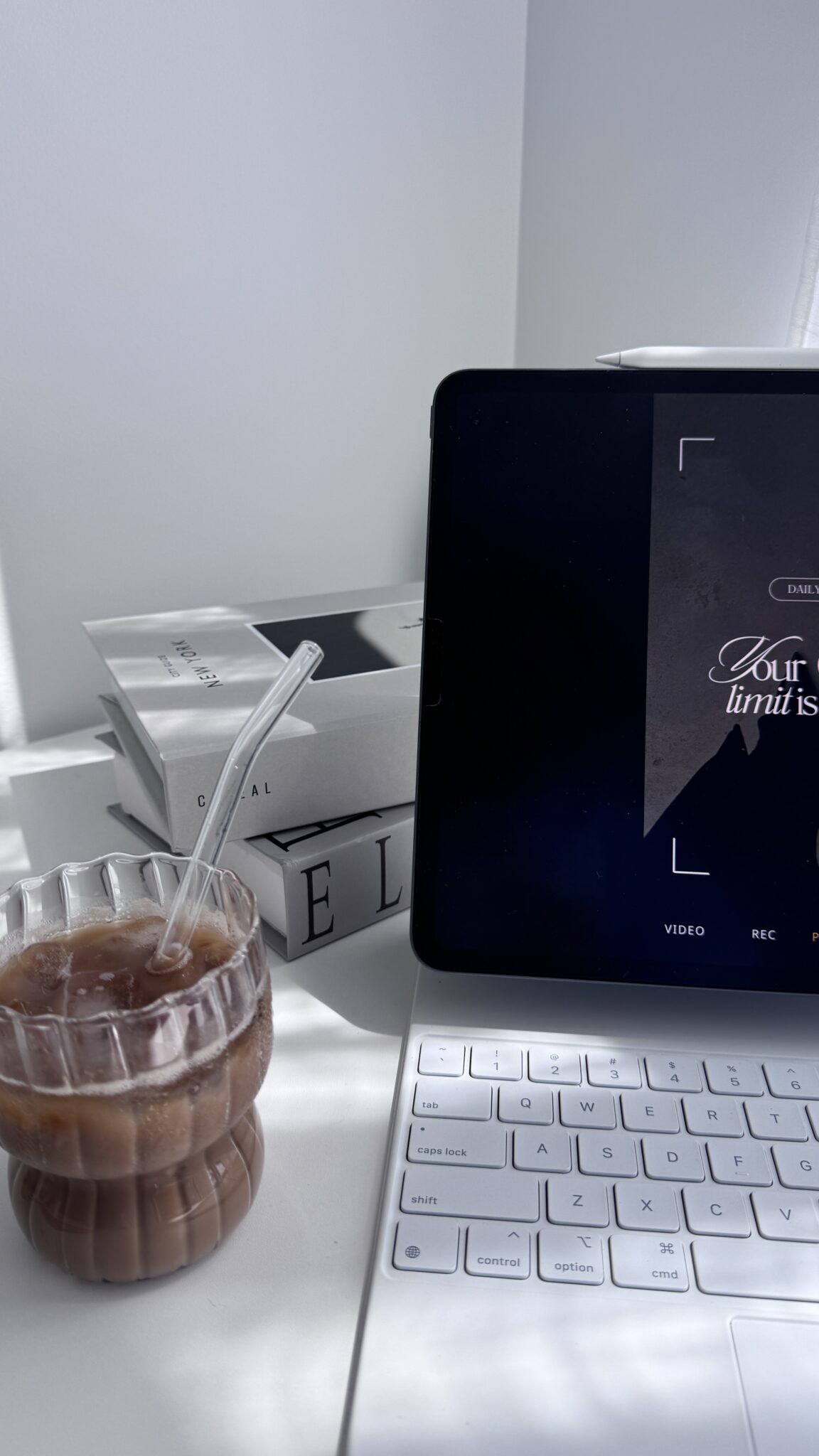 A stylish setup featuring a glass of iced coffee, a tablet displaying a motivational quote, and stacked books in a minimalist workspace.