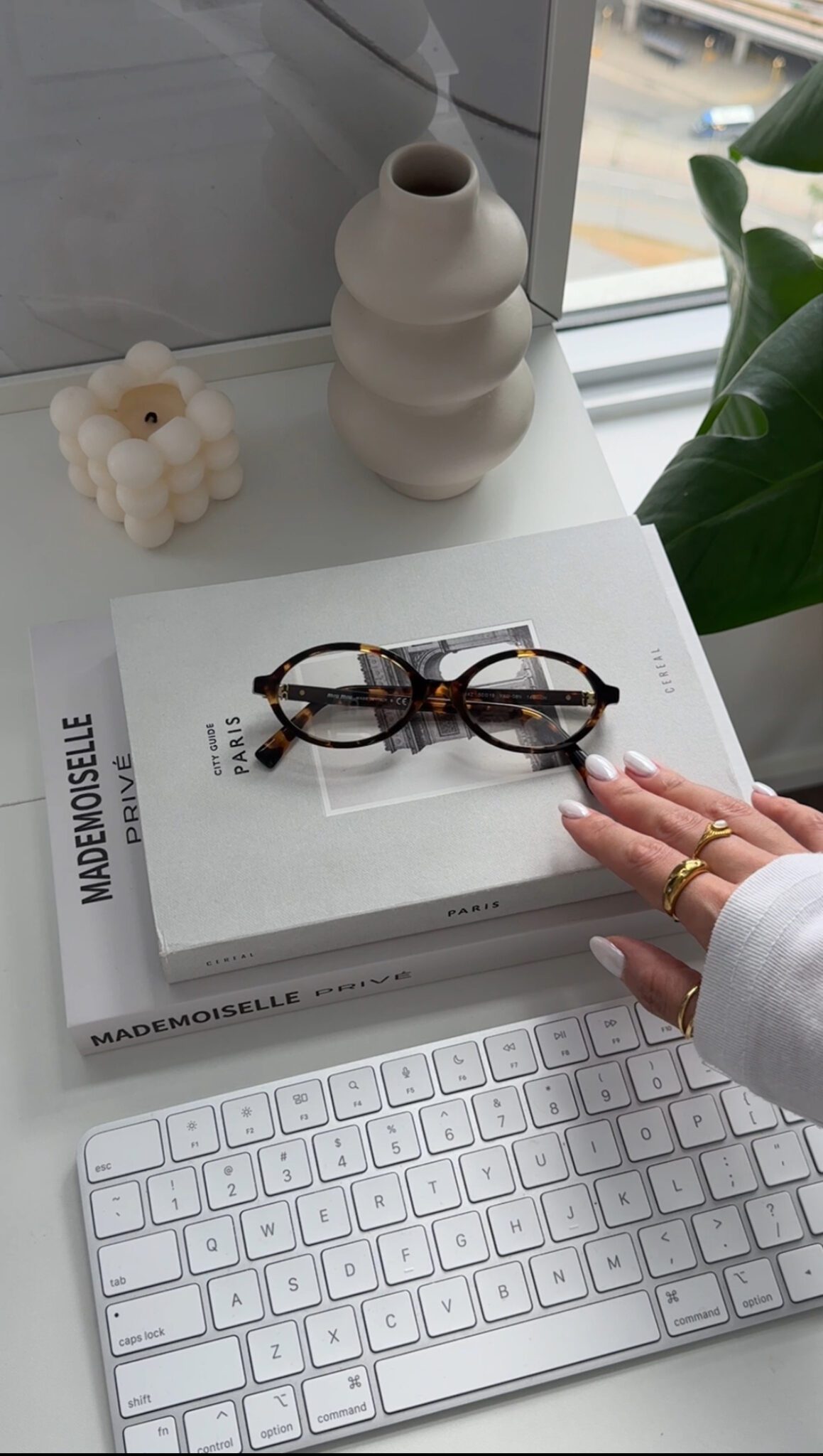 A hand with gold rings reaches for glasses on stacked books near a keyboard and decor items by a window, evoking the aesthetic of Faceless Content Banks.