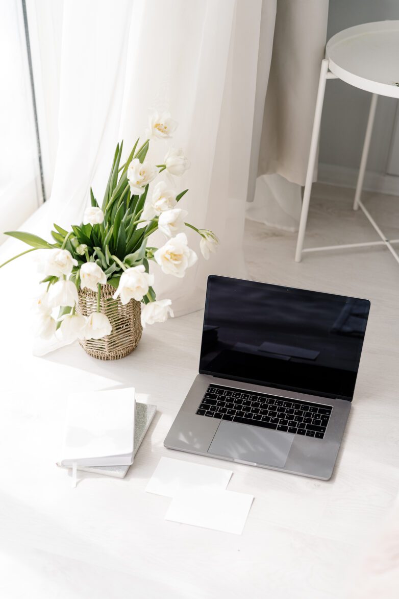 Silver laptop, white tulips in a basket, notebooks, and papers on a bright, minimalist workspace floor—perfect for brainstorming your next faceless marketing strategy.