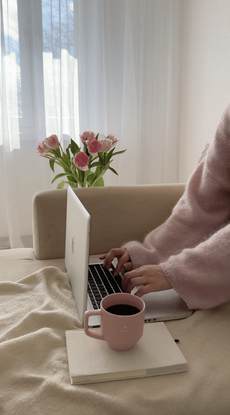 Faceless Content Idea: Person in a pink sweater typing on a laptop with a coffee cup and pink flowers nearby on a beige sofa, creating cozy vibes without showing their face.