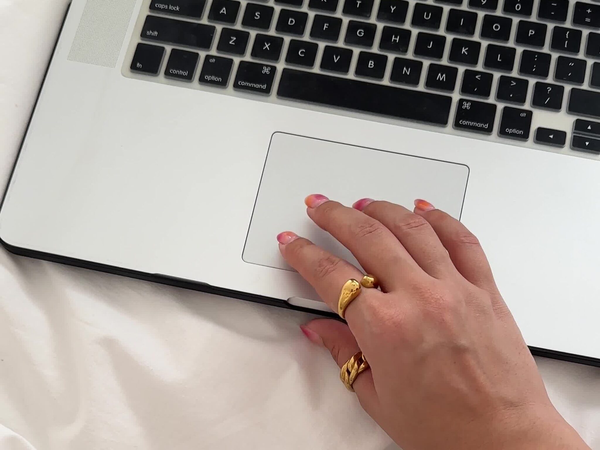 A hand with gold rings rests on a laptop trackpad as someone researches how to start an anonymous blog for free, sitting cross-legged on a bed with white sheets.
