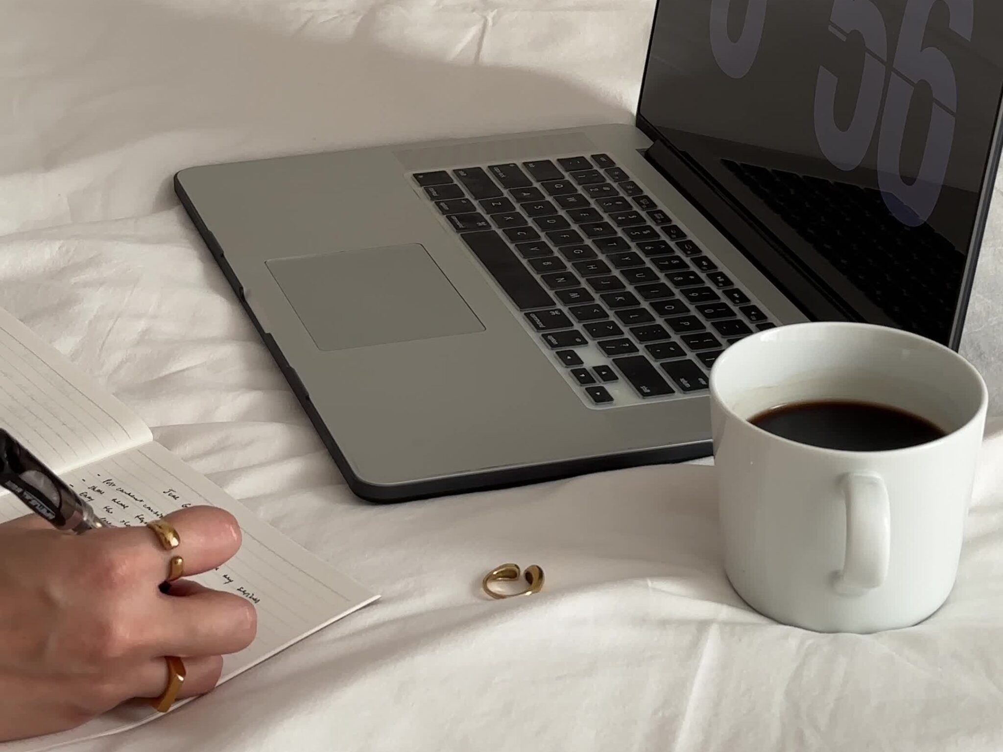 A laptop, cup of coffee, and notebook on a white bed; perfect for researching how to start an anonymous blog for free.