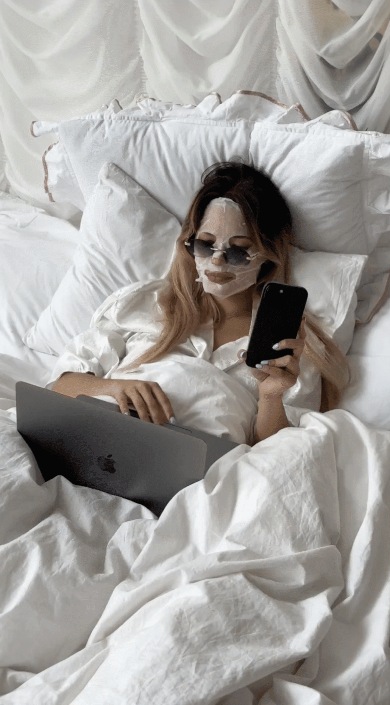 Woman in bed wearing a face mask and sunglasses, using a laptop and holding a phone—a perfect faceless content idea for those seeking privacy and creativity.