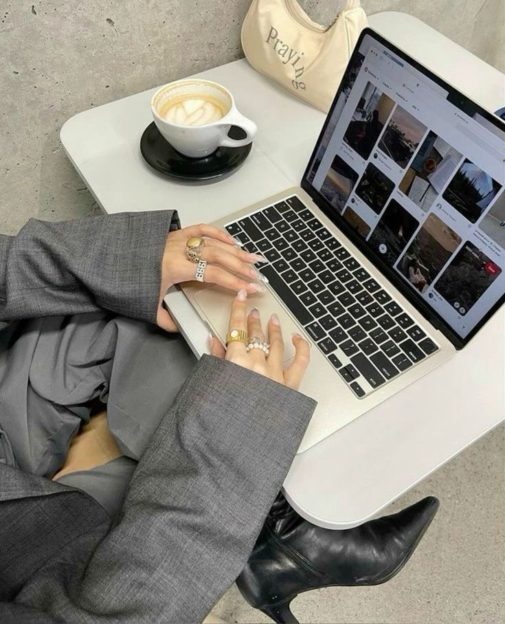 Person in gray suit browsing images on a laptop at a café table with coffee and a small beige bag.