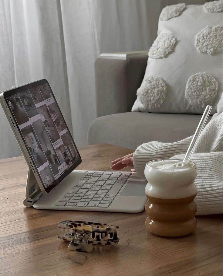 A person using a tablet with a keyboard, a frothy drink, and a hair clip on a wooden table nearby—an ideal setup for crafting a faceless marketing strategy.