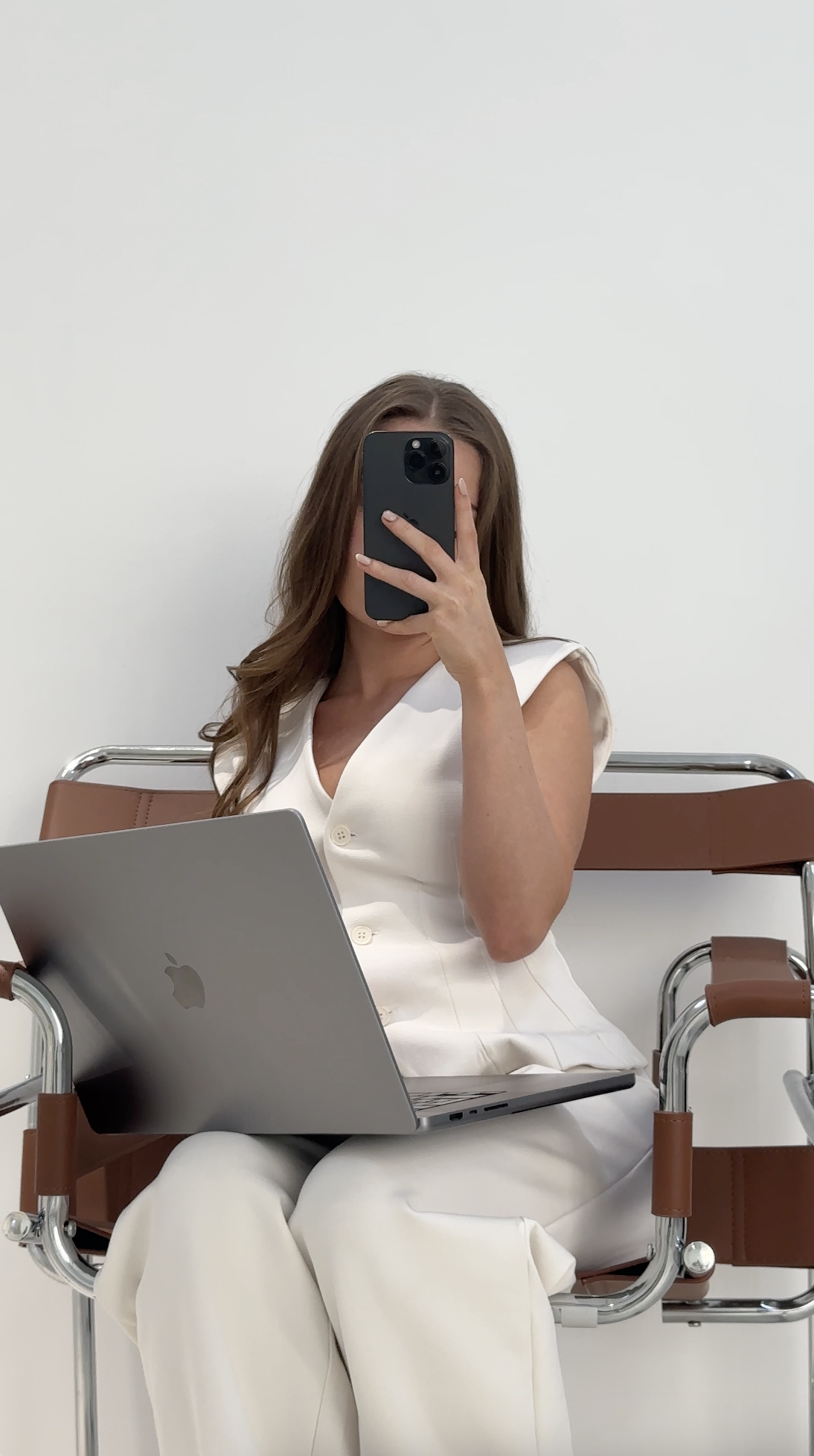 Woman in white outfit sits on a chair, holding a phone and a laptop in her lap, facing a white wall—capturing the essence of How to Create Faceless Content.