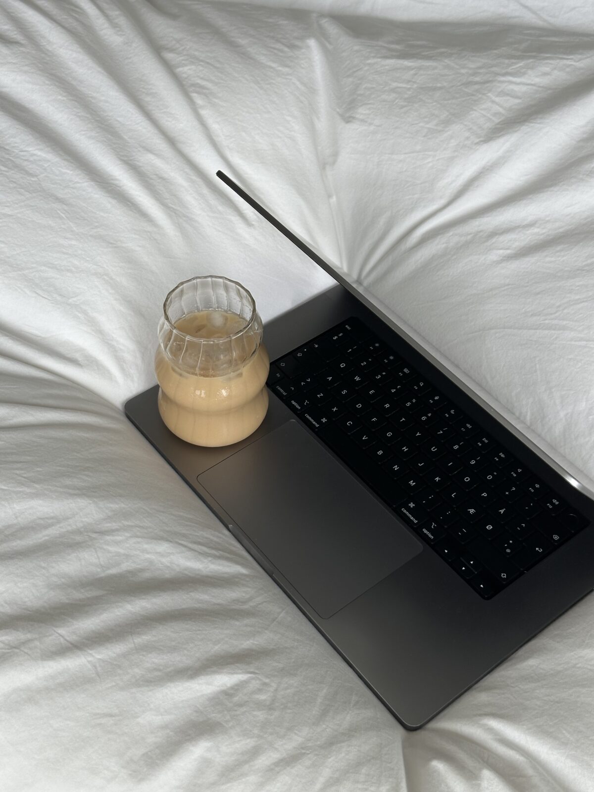 A glass of iced coffee sits on a laptop keyboard atop white, crumpled bedsheets—perfect for a morning spent exploring passive income or planning your next faceless marketing strategy.