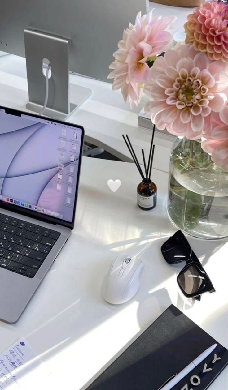 Laptop, mouse, sunglasses, notebook, and a vase with pink flowers on a bright, tidy desk.