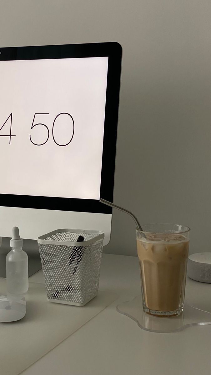 A glass of iced coffee with a metal straw sits beside a computer monitor and desk items on a white desk.