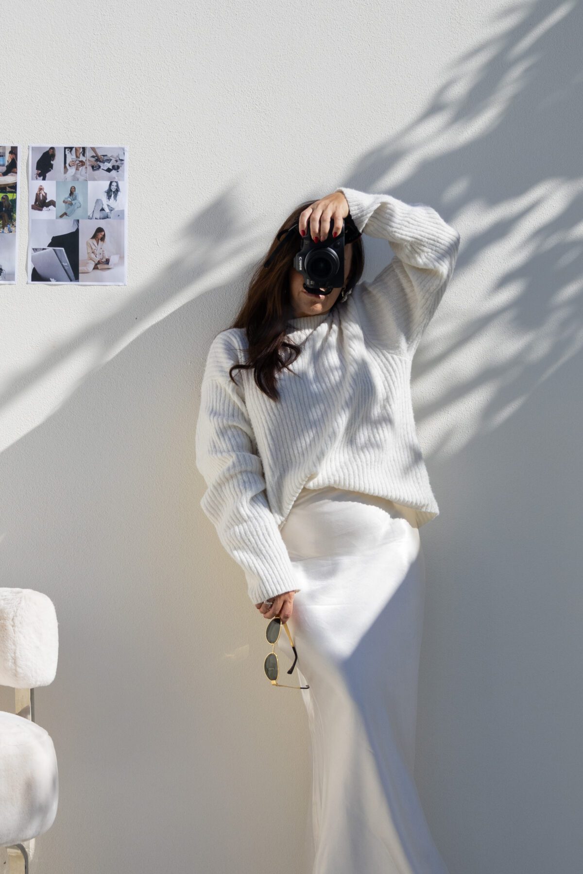 Woman in white sweater and skirt takes a photo with her camera, standing by a white wall with photos and shadows—capturing moments ideal for showcasing the easiest Faceless Business ideas.