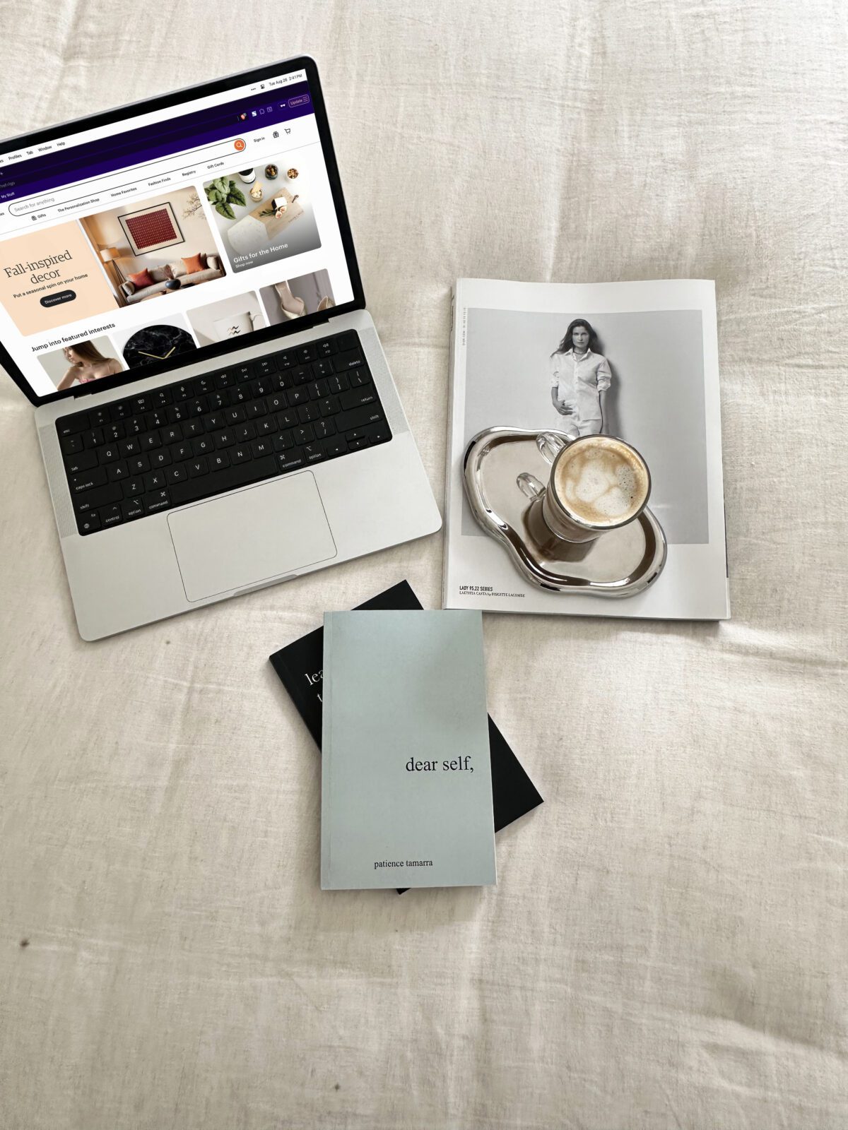 A laptop, coffee, magazine, and books are arranged on a beige fabric surface—perfect essentials for planning your easiest Faceless Business.