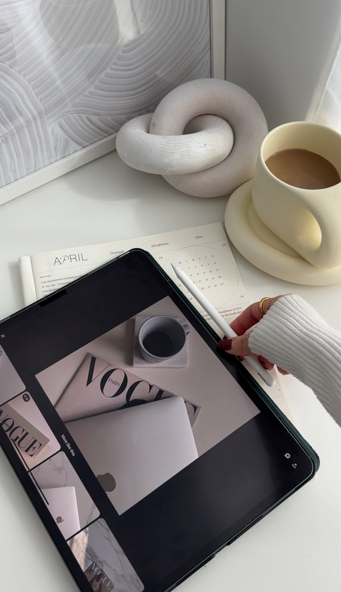A hand uses a stylus on a tablet near a coffee cup, calendar, and abstract decor—perfect for creating faceless content banks in a modern workspace.
