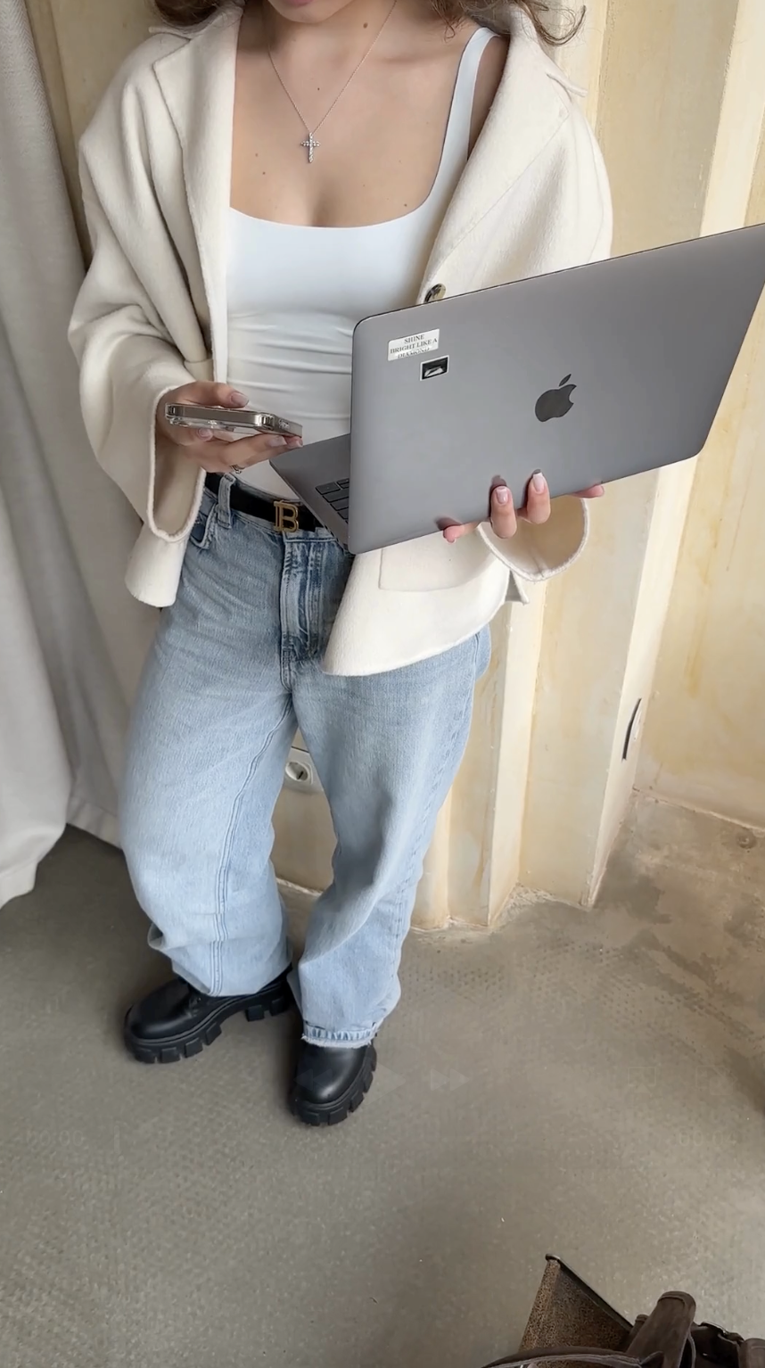 Person in jeans and a white jacket holds a laptop and phone while standing indoors on a concrete floor, perhaps researching What Is Faceless Marketing.