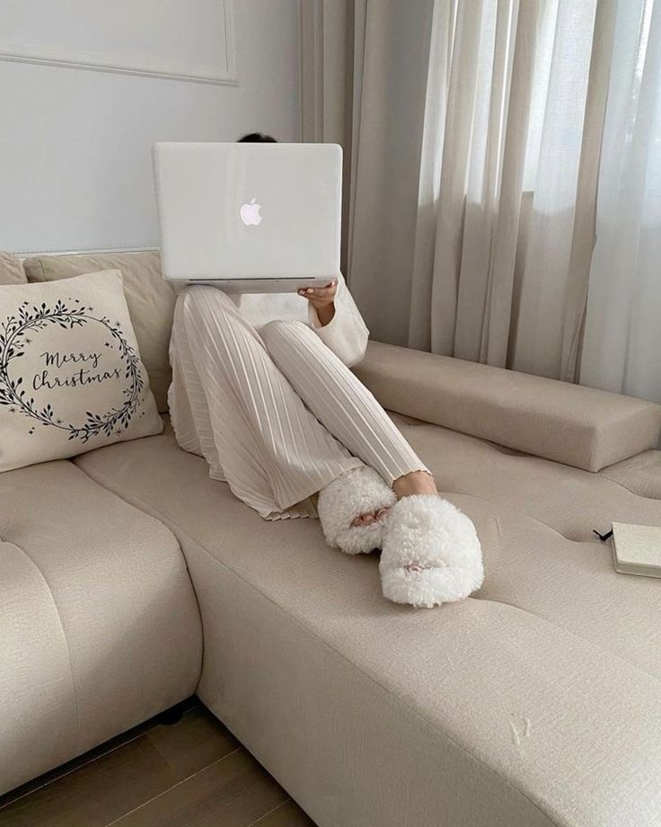 Person in cozy loungewear and fluffy slippers sits on beige sofa using a laptop, Merry Christmas pillow nearby—capturing the essence of a faceless marketing strategy with relatable, inviting comfort.