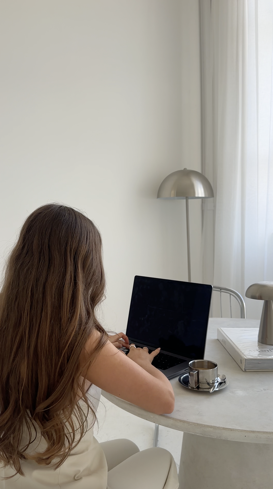 A woman with long brown hair works on Faceless Affiliate Marketing at a white desk with coffee and books in a bright, airy room.