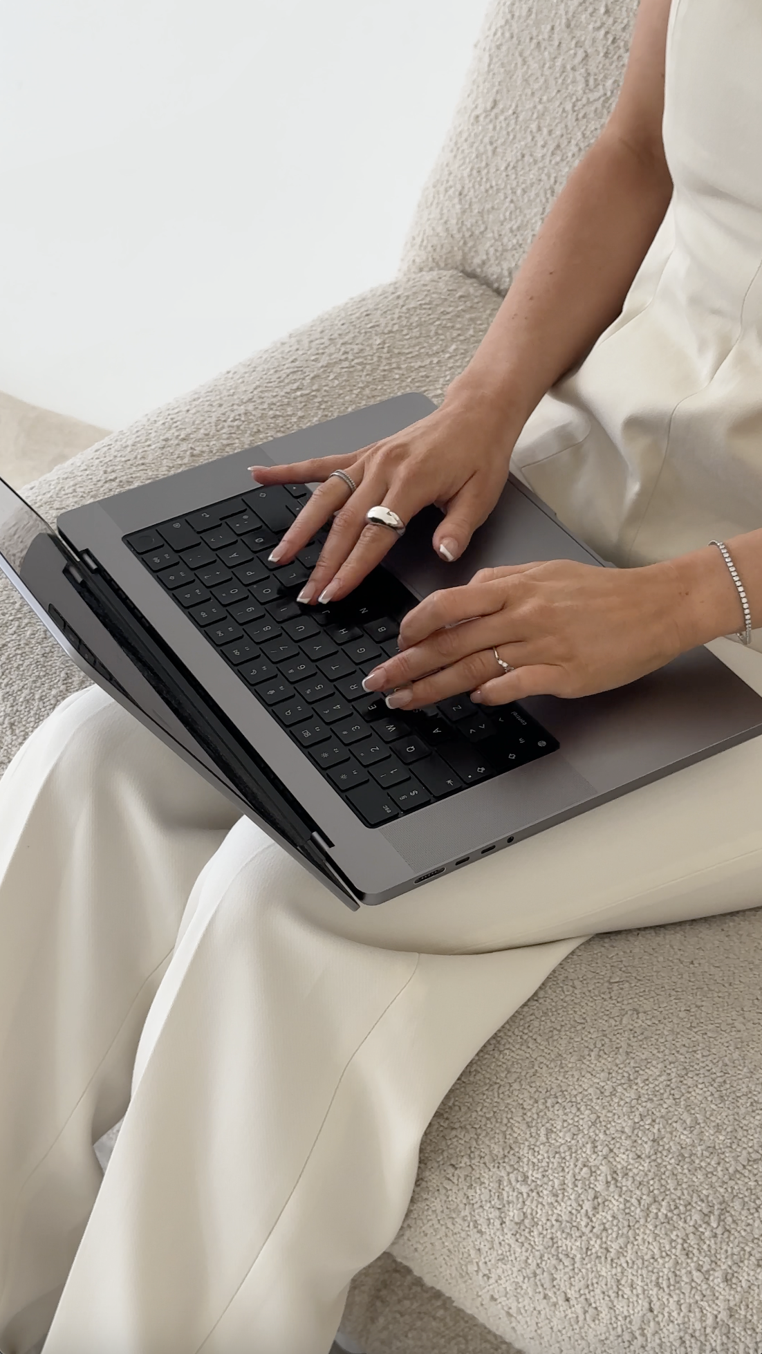 A person in white clothing types on a laptop while sitting on a textured light-colored chair, embodying the quiet focus of building passive income through faceless marketing.
