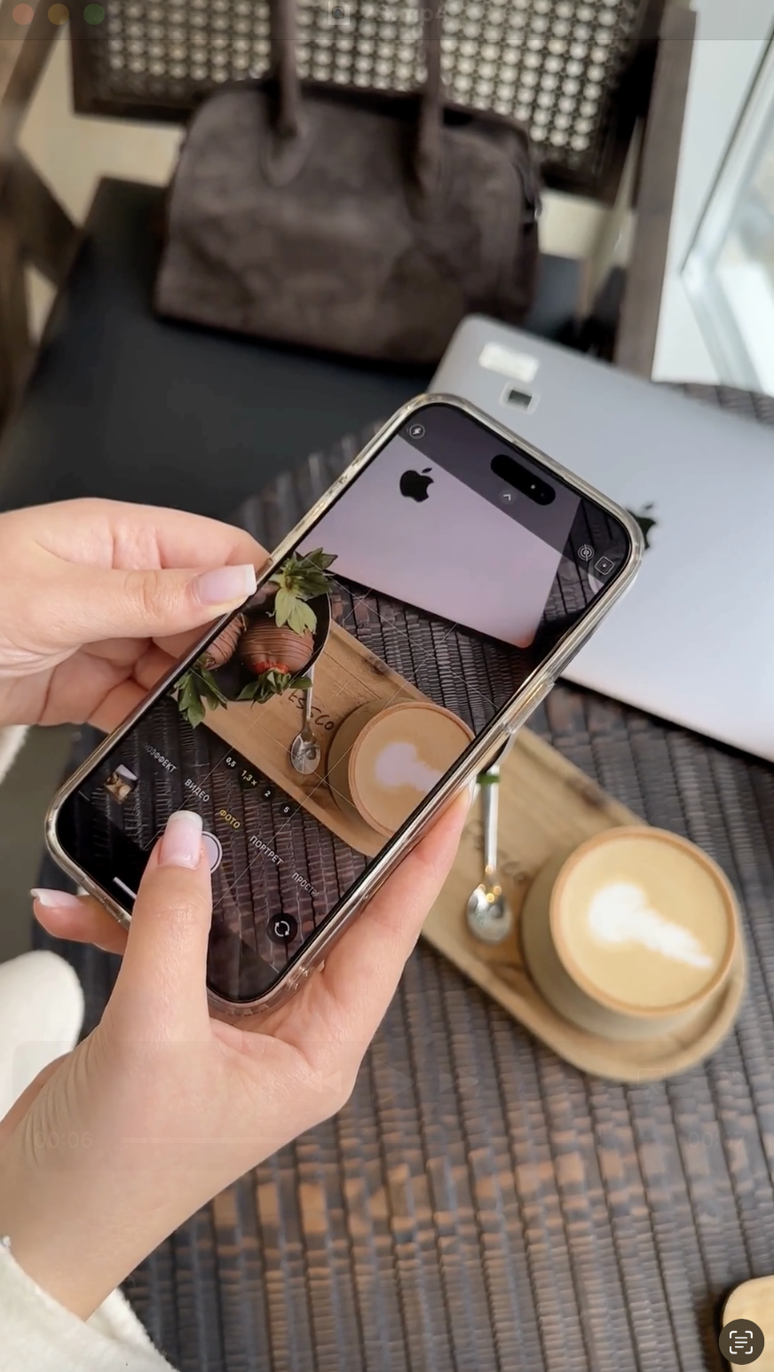 A person holds an iPhone photographing a latte and dessert on a wooden tray next to a laptop, capturing content perfect for exploring What Is Faceless Marketing.