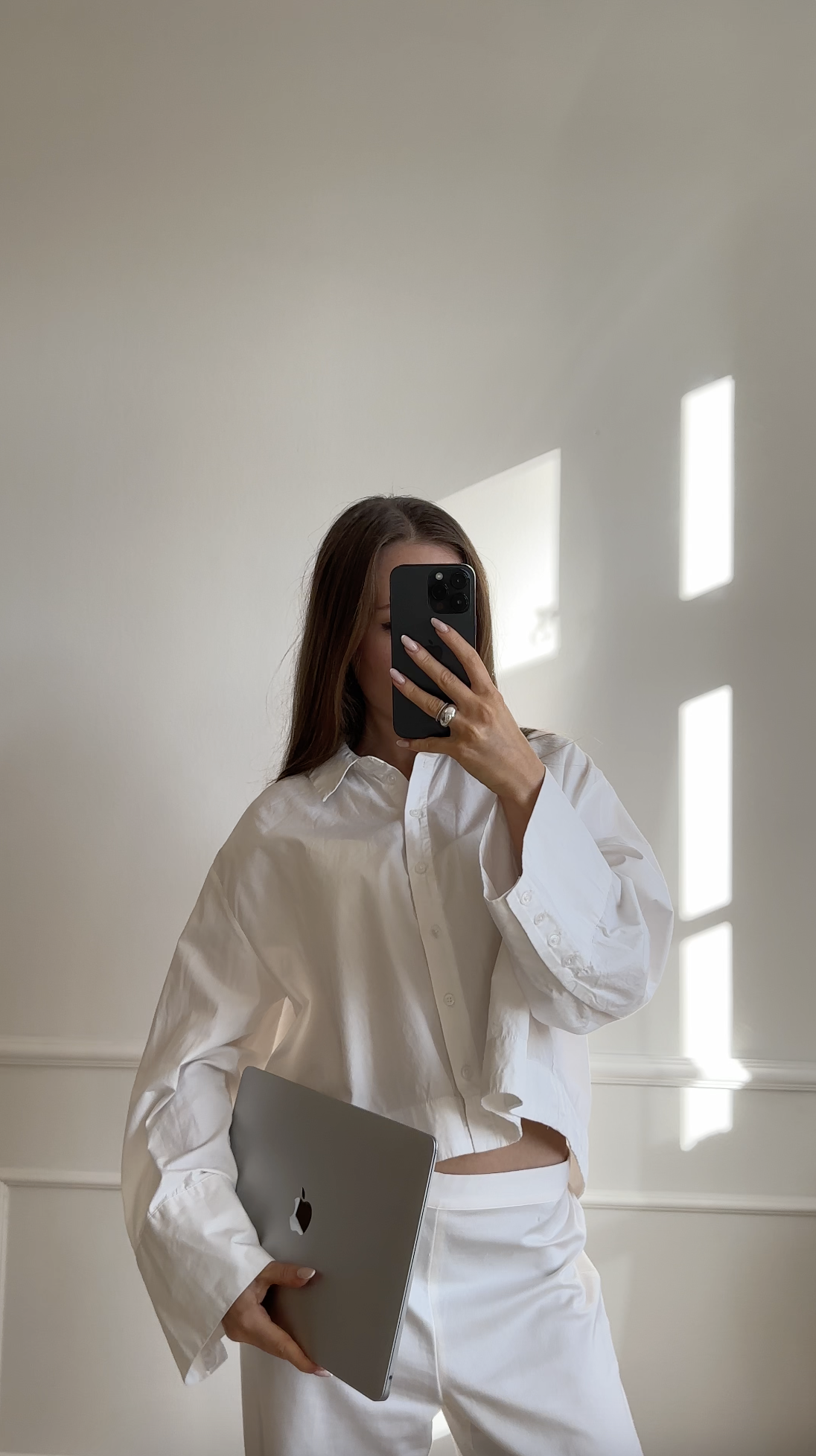Woman in white shirt holding a laptop and taking a mirror selfie with a phone in a sunlit room—capturing the essence of Faceless Affiliate Marketing.