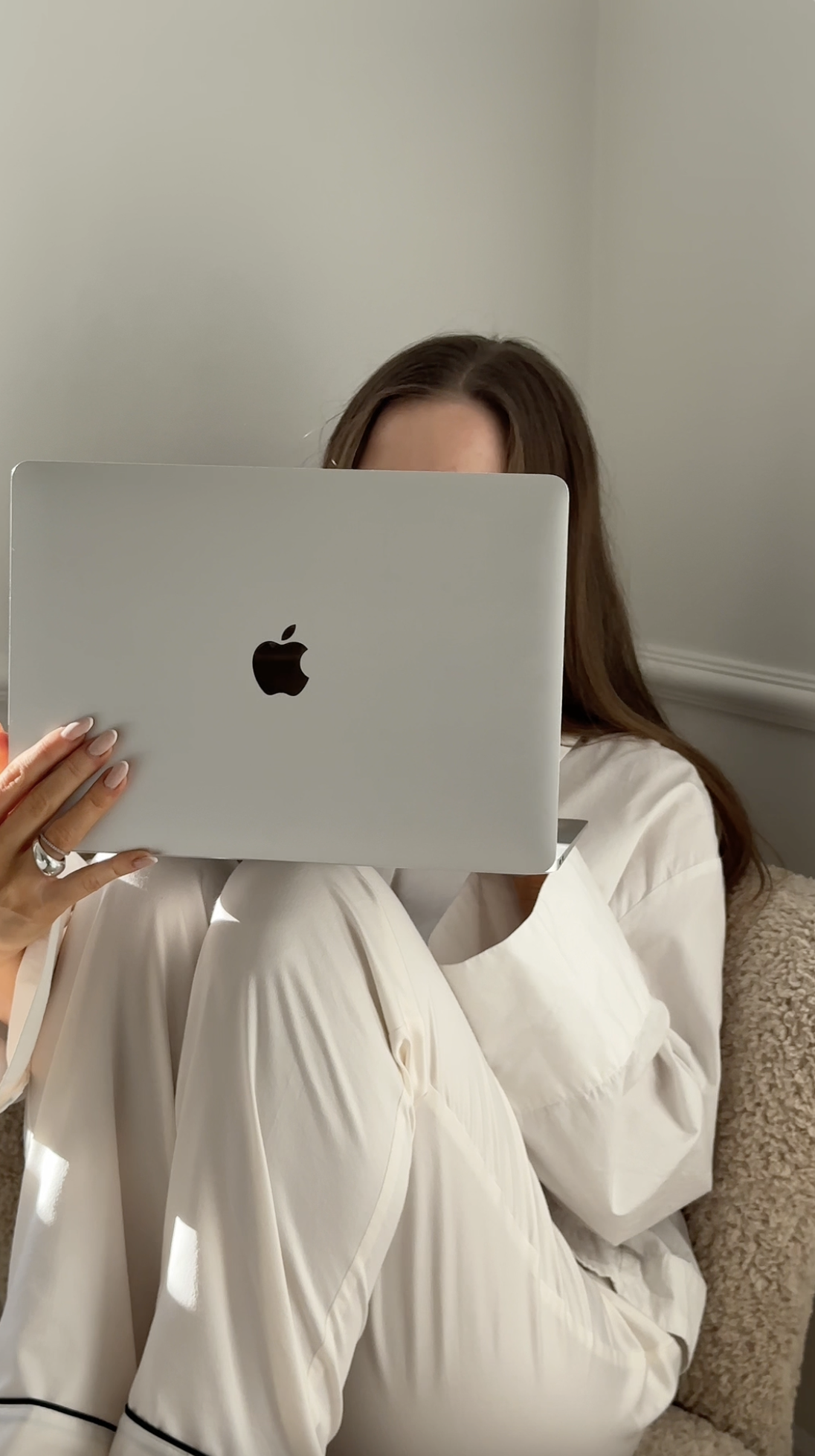 A person in white clothes sits with knees up, holding an open MacBook laptop in front of their face, perfectly embodying the concept of Faceless Affiliate Marketing.