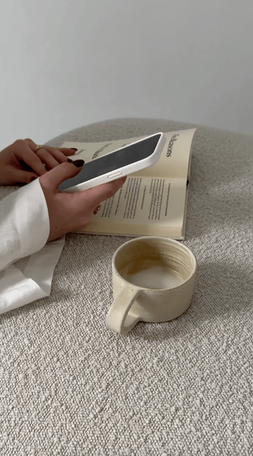 A person holds a phone over an open book beside an empty beige mug on a textured surface.