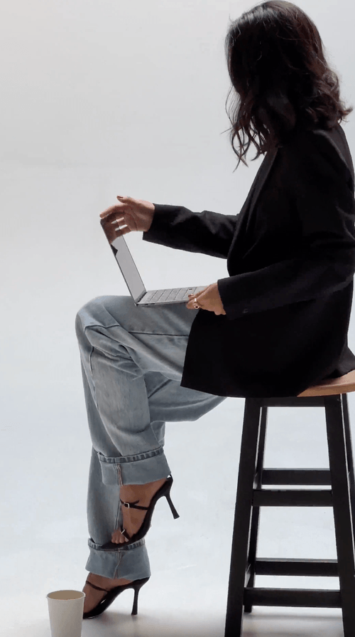 Faceless Content Banks: Woman in jeans and heels sitting on a stool, holding a laptop, with a coffee cup on the floor—perfect for faceless content banks seeking modern, versatile imagery.