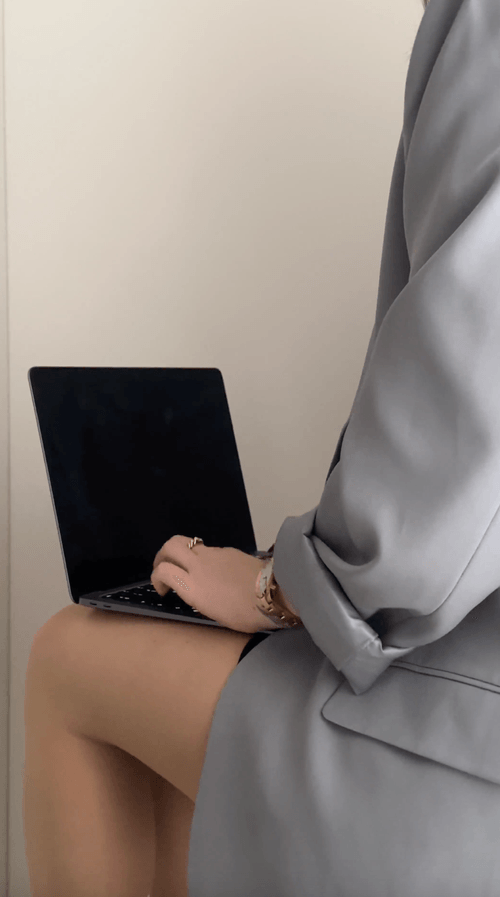 A person in a gray blazer sits with a laptop on their lap, typing with one hand, embodying the rise of faceless business ideas 2025 against a plain background.