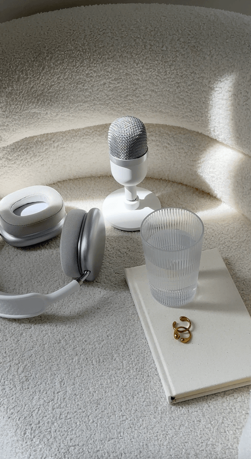 White headphones, a microphone, a glass of water, a notebook, and a ring on a textured white surface.