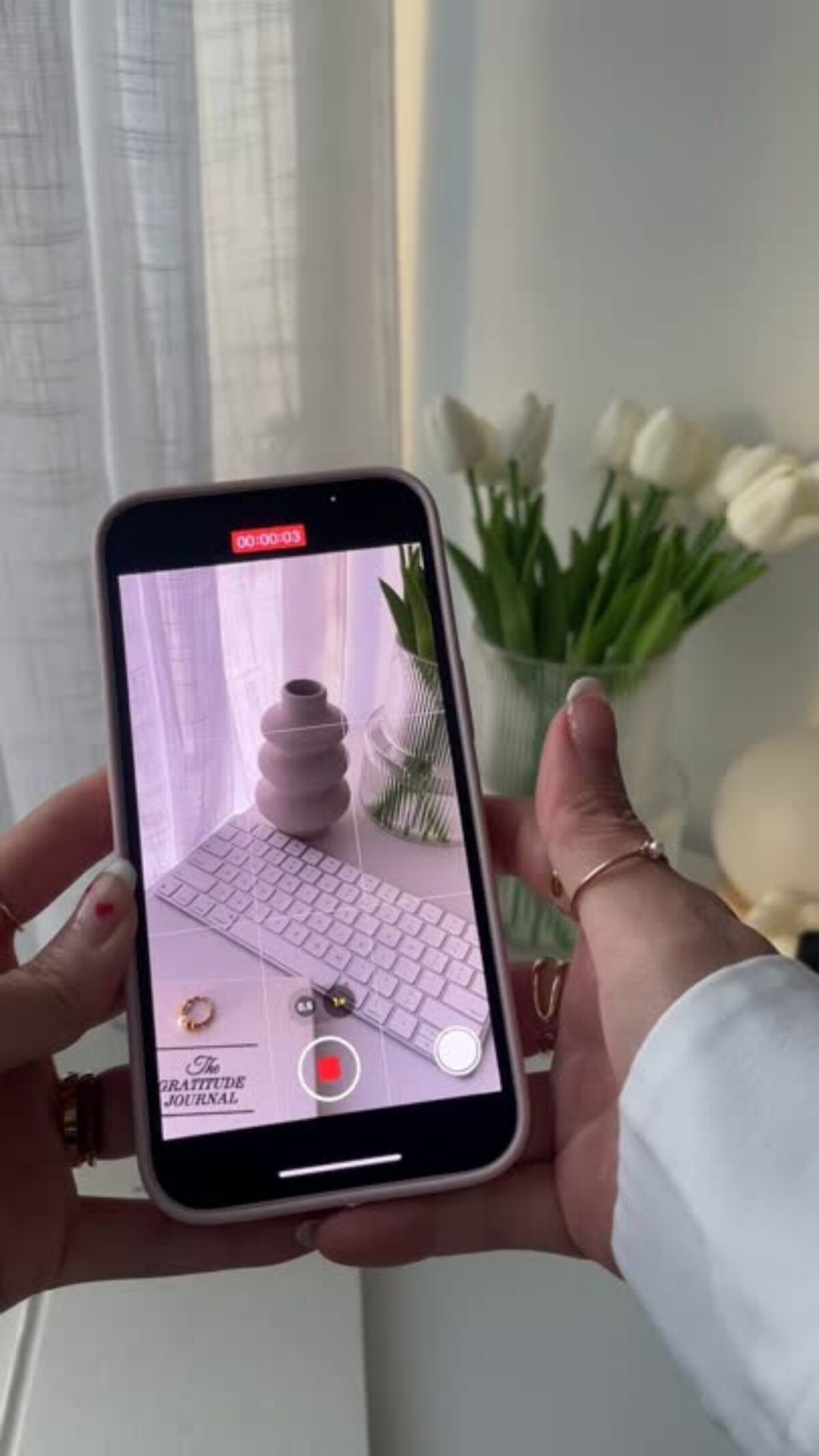 A hand holds a phone filming a white keyboard and vase, with tulips and decor in the background—a perfect Faceless Content Idea for capturing minimalist workspace inspiration.