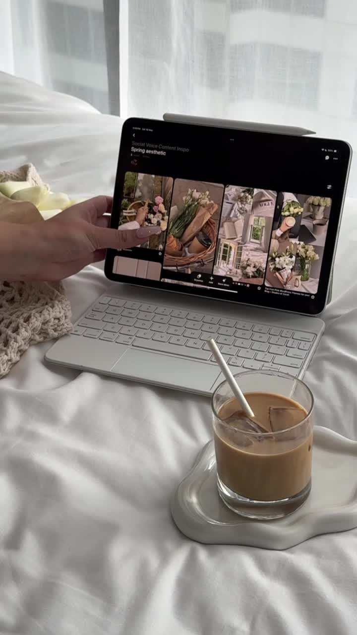 A hand holds a tablet showing flower photos beside iced coffee on a white bed with a keyboard—perfect inspiration for learning how to start faceless digital marketing from the comfort of home.