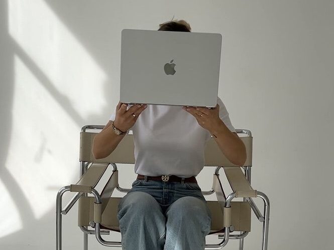 Person sitting on a chair holding a laptop in front of their face, casting soft shadows on a white floor—perfectly capturing the intrigue behind How to Start an Anonymous Blog for Free.